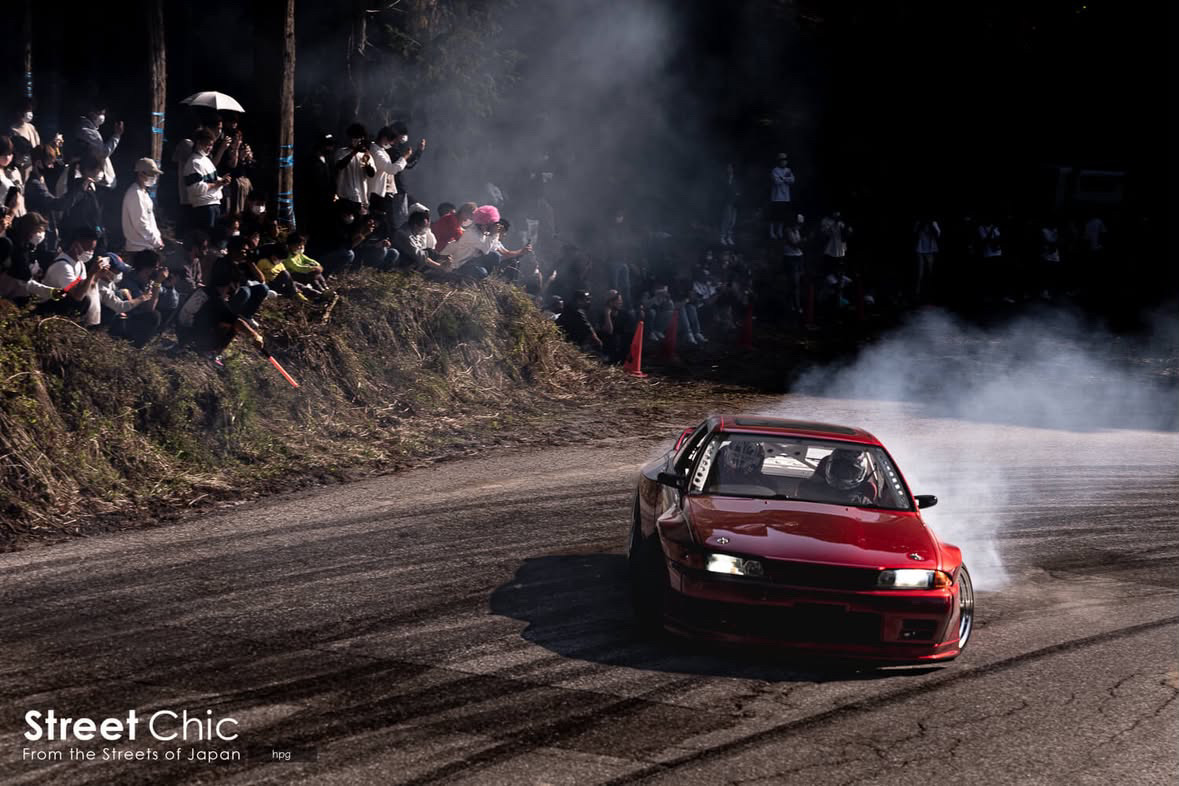 A red Nissan Skyline R32 drifting through tyre smoke past a bank of spectators on a Japanese drift course