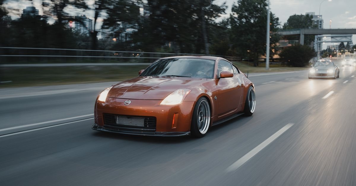 An orange Nissan 350Z with polished wheels driving on a city road at dusk (stock photo, not taken in Japan)