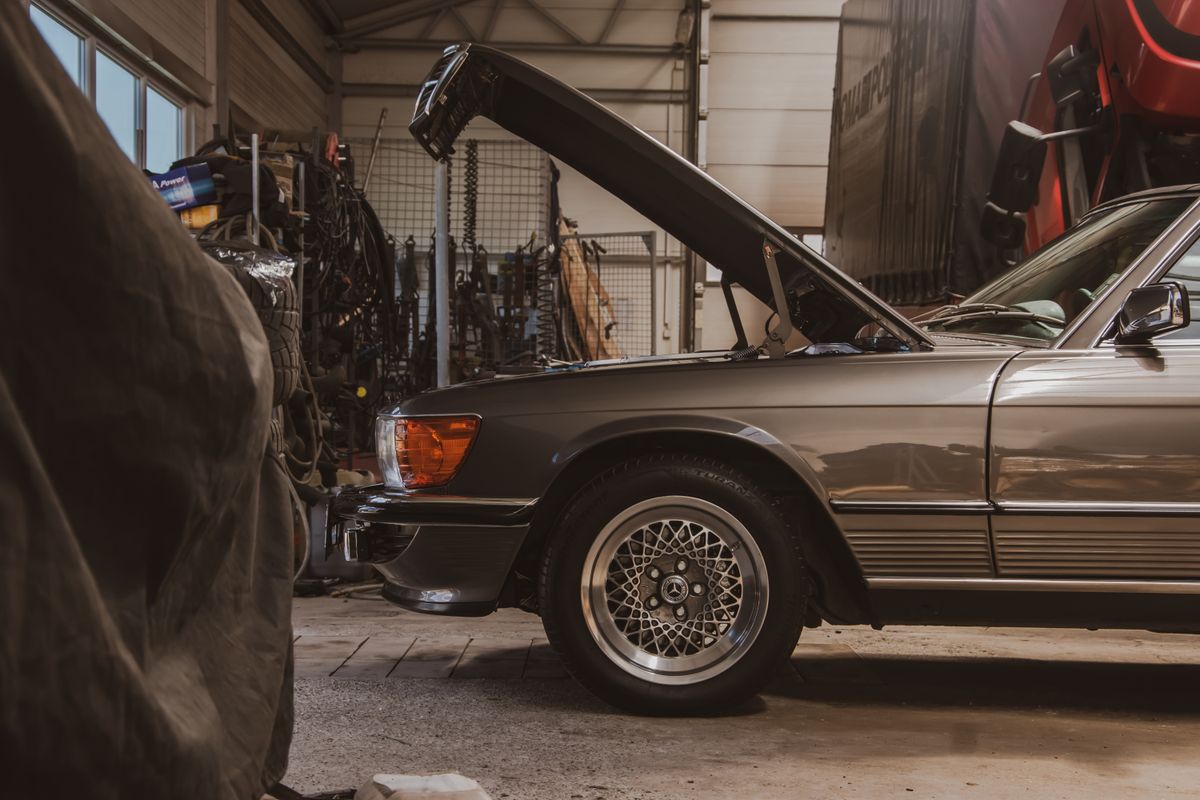 An older coupe kept indoors at a workshop with its bonnet up, another car under a fabric cover in the foreground and parts racks behind