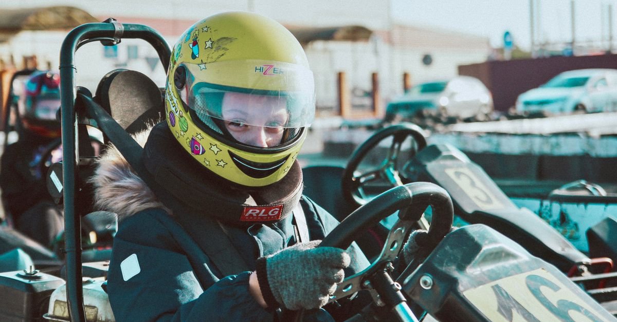 A child in a yellow full-face helmet and neck collar holding the wheel of a go-kart, with other karts parked behind. A kart track, not a drift car, location not identified