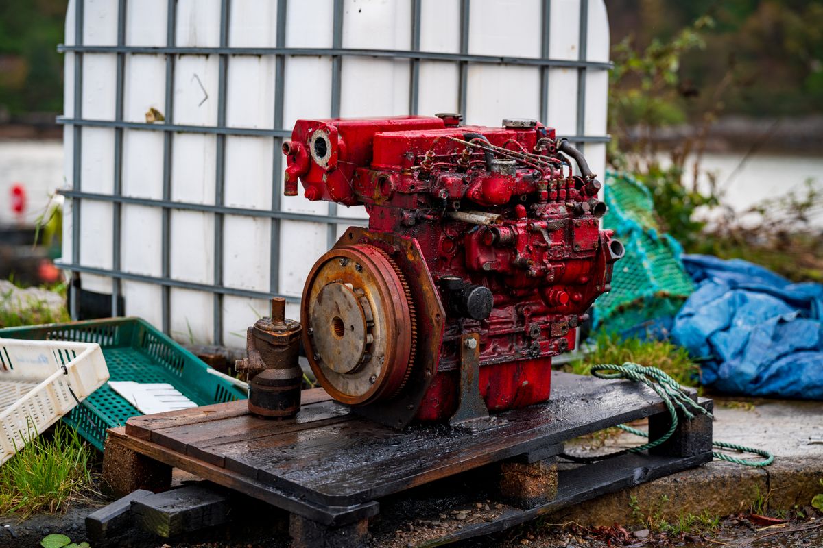 A used engine standing on a wet wooden pallet outdoors, flywheel and bellhousing face turned towards the camera
