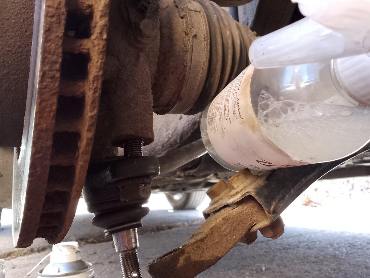 A rusty front steering knuckle with the brake rotor and driveshaft boot attached, its lower ball joint separated from the control arm (an everyday front-wheel-drive car, not a Silvia, Chaser or 350Z)