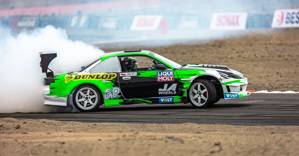 A green and black drift car pouring tyre smoke in front of sponsor banners at a drift competition (stock photo, not taken at Hadashi Tengoku Circuit)