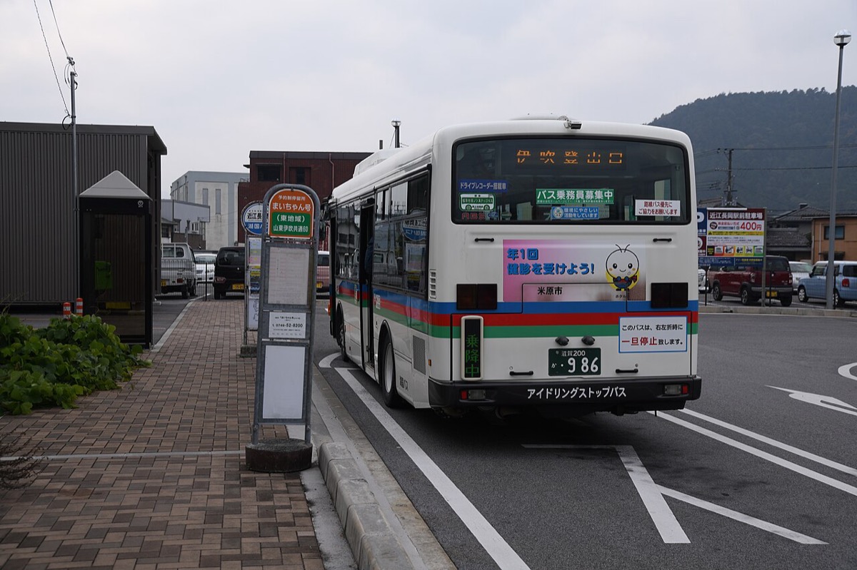 The forecourt of JR Ōmi-Nagaoka Station in Maibara, Shiga: a Maichan-go shared-taxi stop sign beside a Kokoku Bus bound for Ibuki-tozanguchi, not for Okuibuki