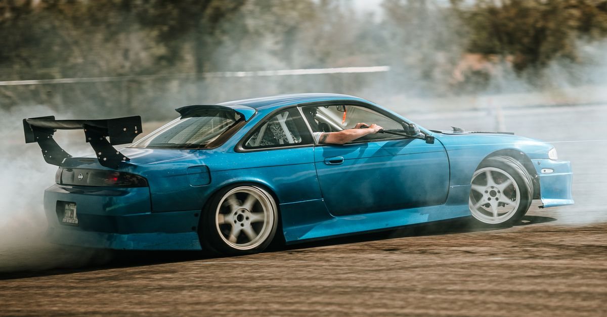 A blue Nissan Silvia S14 sliding sideways in tyre smoke on a dry outdoor course
