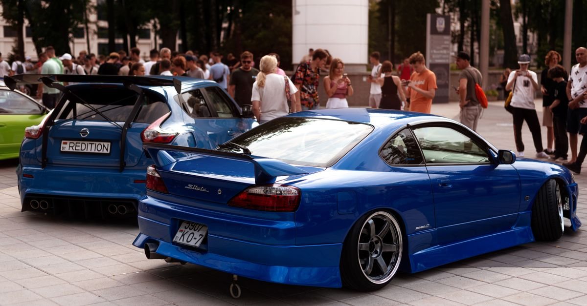 A blue Nissan Silvia S15 with a boot-lid rear wing parked next to a blue hatchback with a tall GT wing at an outdoor car meet. Stock photo, not ORIGIN Labo. parts