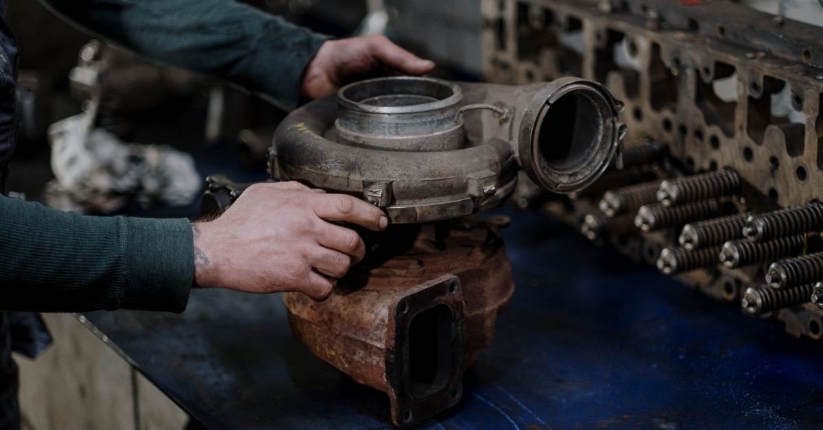 Hands holding a used, rust-stained turbocharger on a workshop bench next to a stripped cylinder head