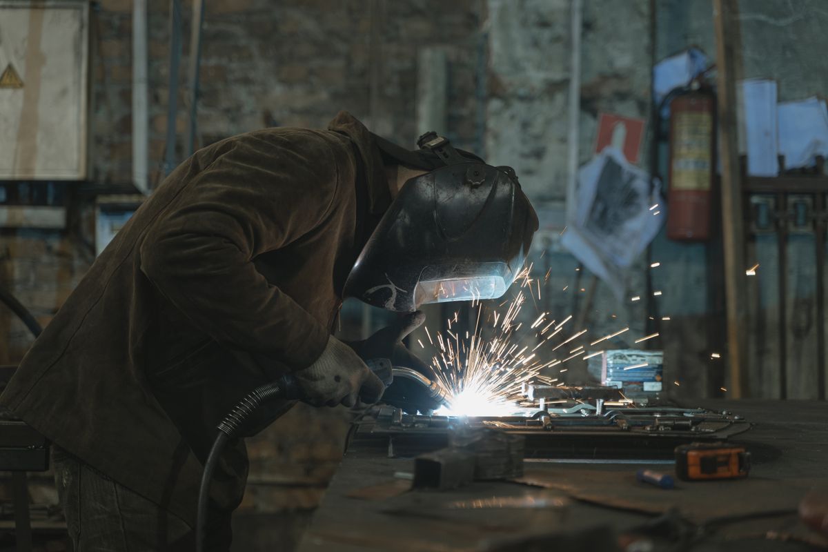 A fabricator in a leather jacket and welding mask MIG-welding a steel part on a workbench, sparks flying